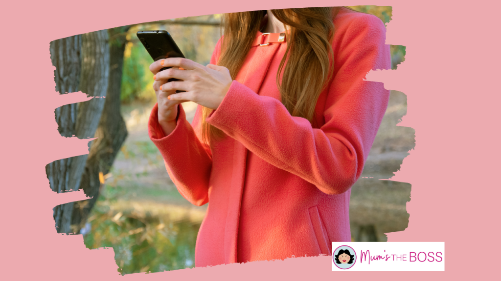A woman is looking at her phone while standing in a forest. She is wearing a fashionable orange jacket but the photo does not show her face